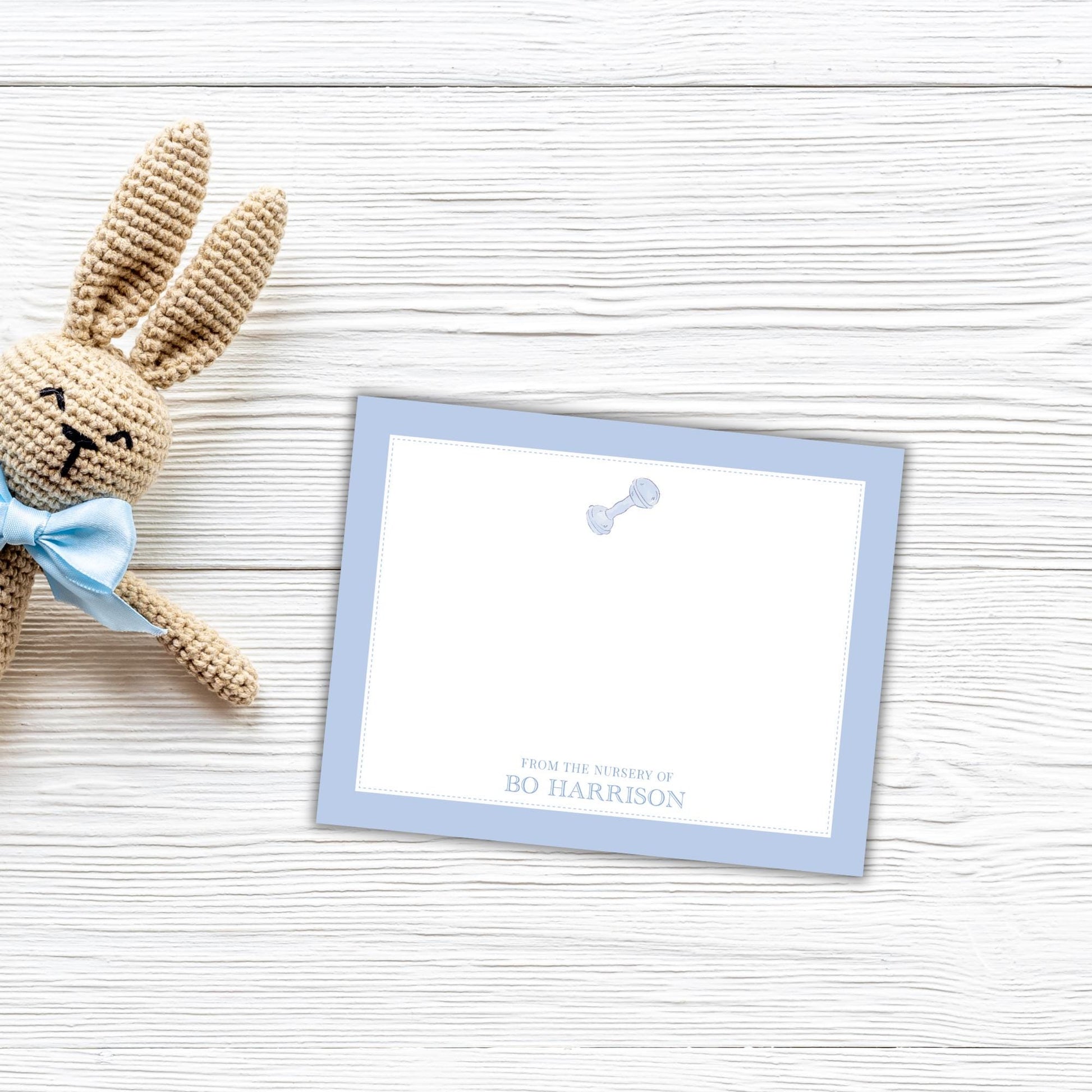 A crocheted bunny toy is placed next to a blank note card with a personalized message on a white wooden surface.