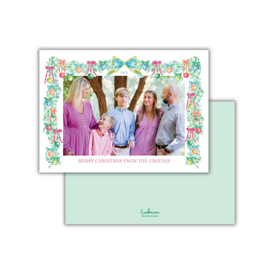 A family portrait of a group of people, likely a family, posing together in front of a Christmas tree. The family members are dressed in colorful holiday attire, and the background features a festive tree decorated with ornaments and ribbons. The card has a green border and a message that reads &quot;Merry Christmas from the Greens&quot;.