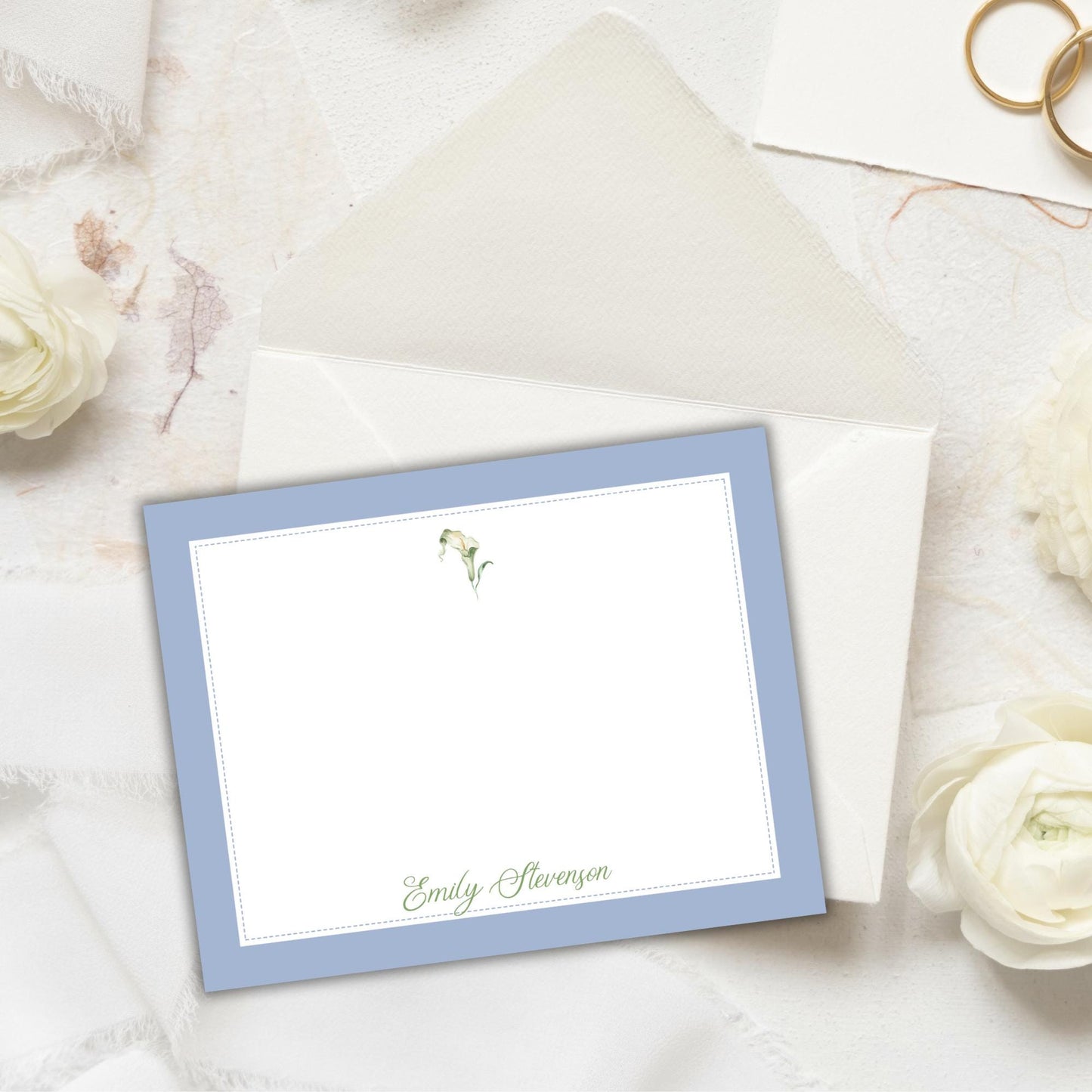 a blue-bordered personalized note card with a name printed on it, placed on a white surface with white flowers and a gold ring nearby.