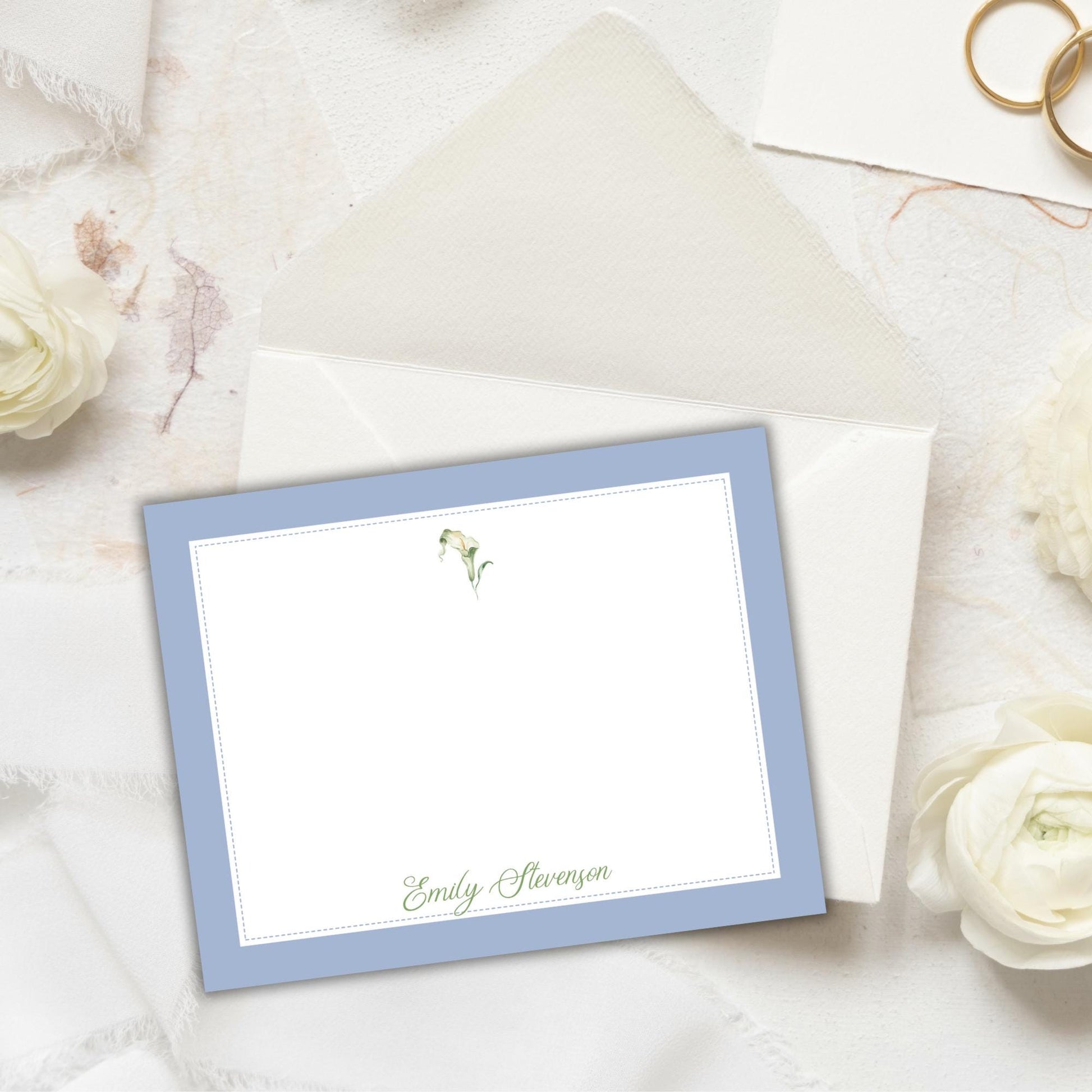 a blue-bordered personalized note card with a name printed on it, placed on a white surface with white flowers and a gold ring nearby.