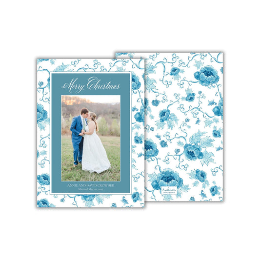 A wedding invitation card with a couple standing in a field, surrounded by a floral pattern in shades of blue.
