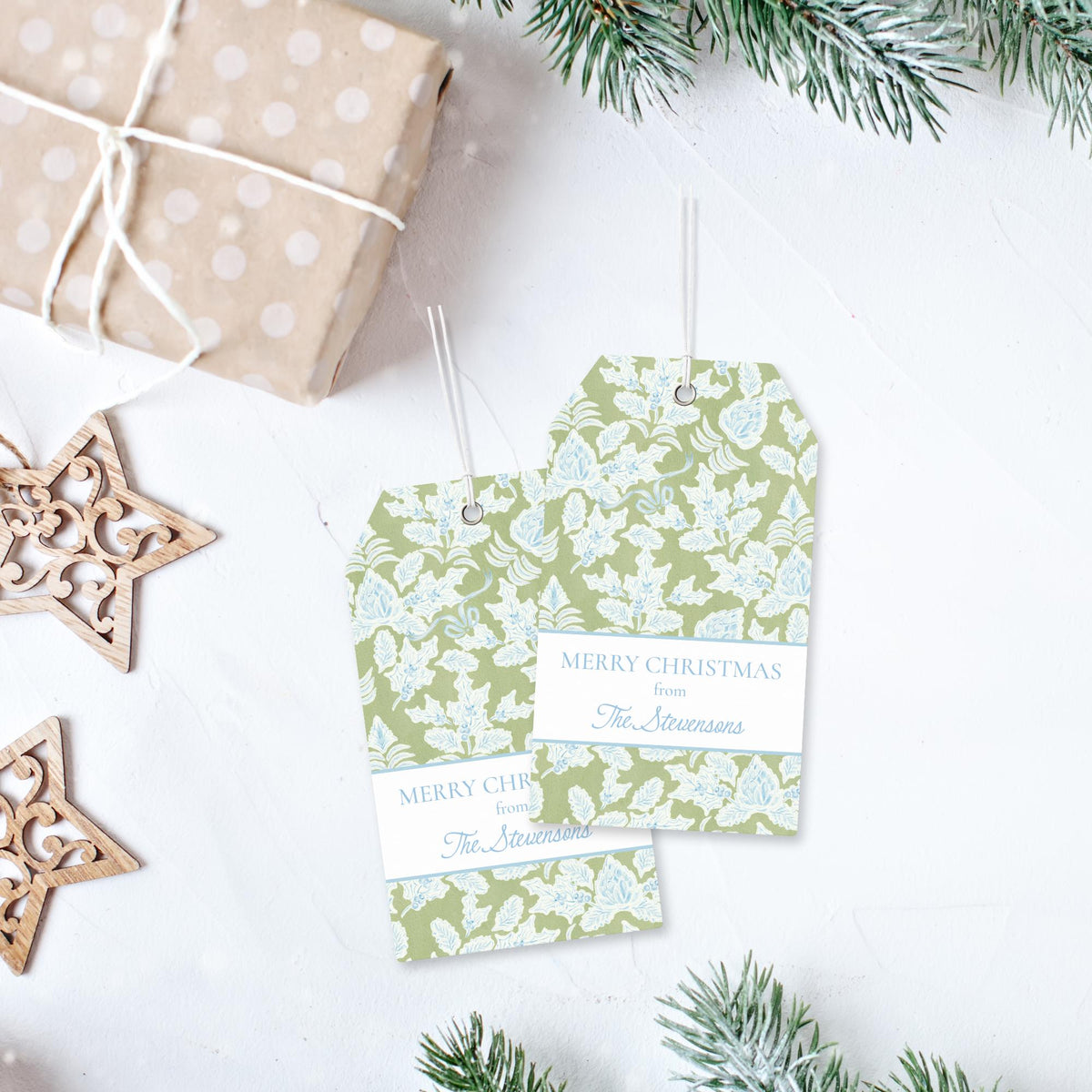 two green and white floral-patterned gift tags with the text "Merry Christmas from The Stevens" printed on them, placed on a white surface next to a gift box and some pine branches.