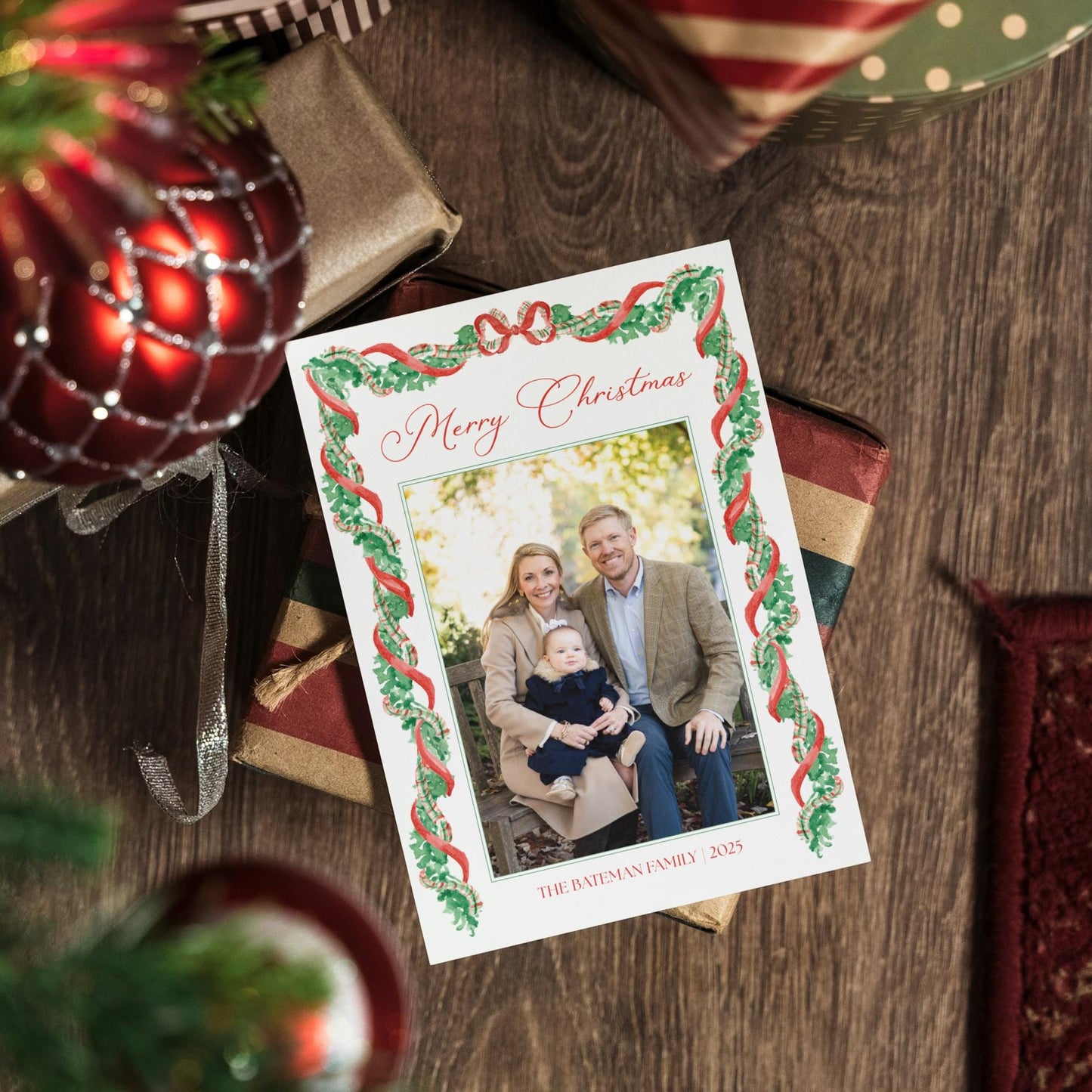 A Christmas card with a family photo is placed on a wooden surface, surrounded by holiday decorations such as ornaments and wrapped gifts.