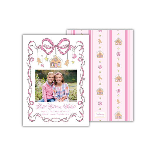 A Christmas card featuring two young girls sitting together in a garden, with a bow and ribbon design around the edges. The card has a pink and white striped background with illustrations of houses and stars.