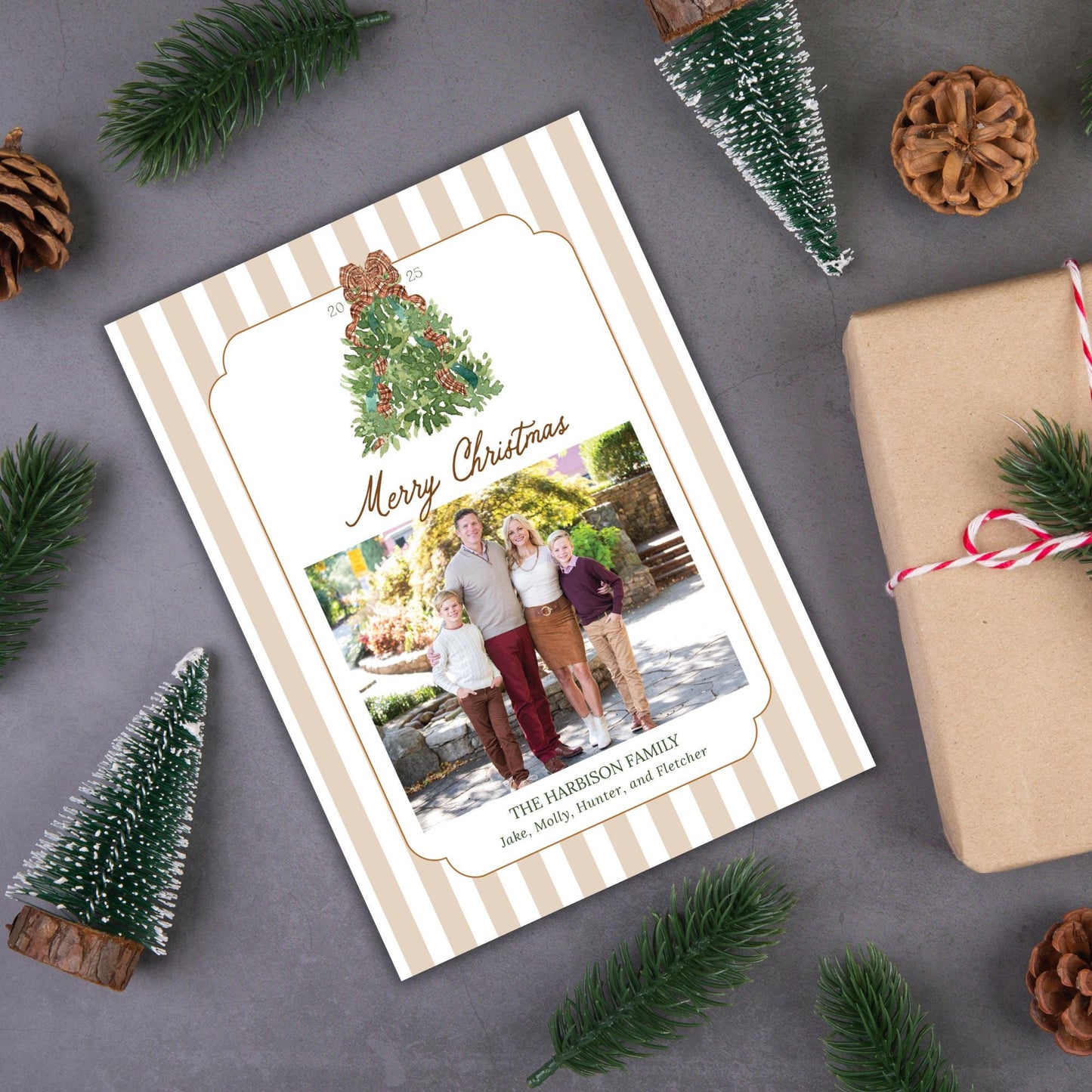 A Christmas card with a family photo and the text &quot;Merry Christmas&quot; is surrounded by pine branches, pine cones, and a gift box on a gray surface.