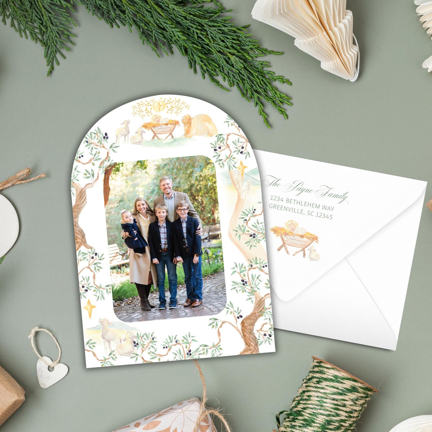 A family portrait is displayed on a white card with a floral border, surrounded by holiday decorations such as pine branches, a ribbon, and a small white envelope.