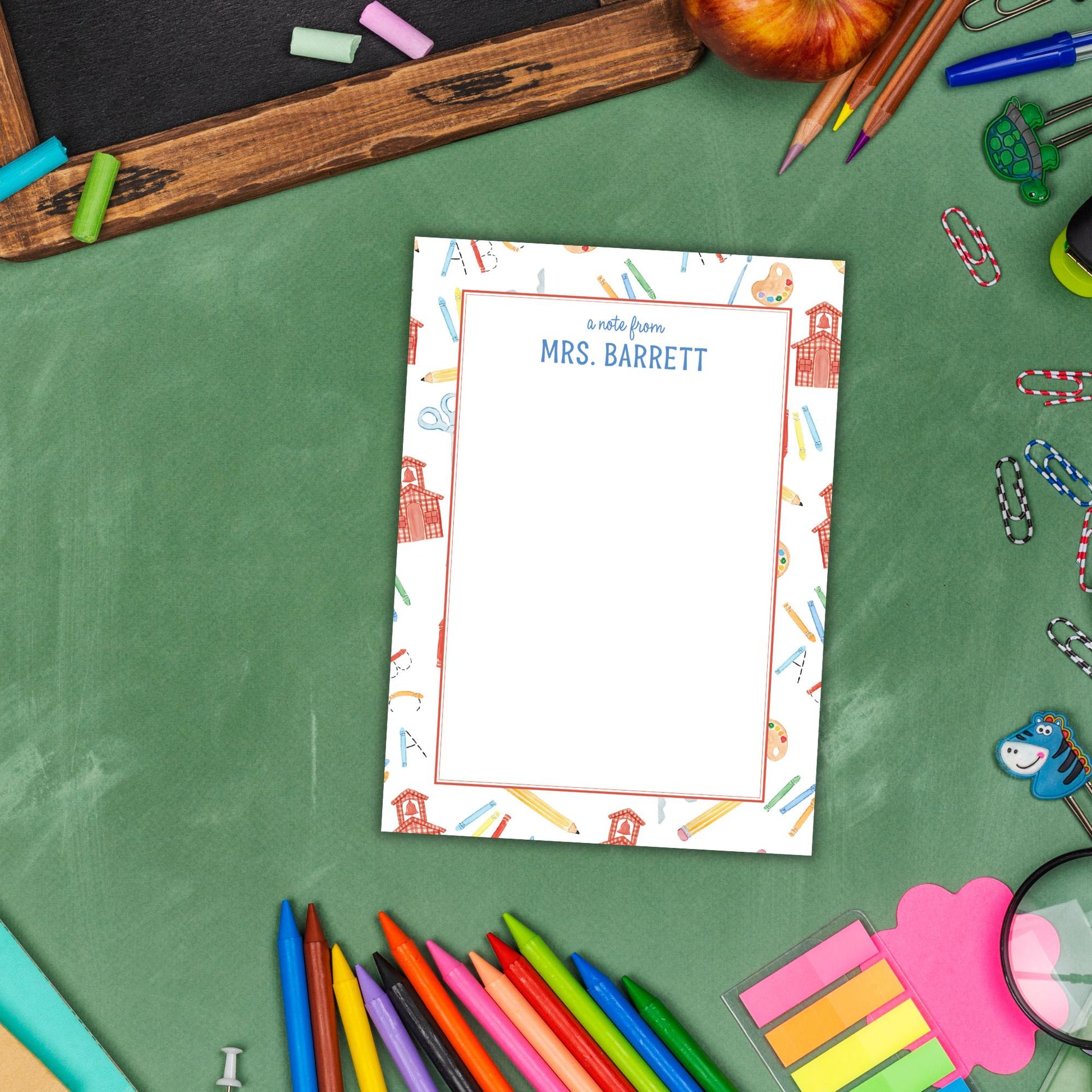 A blank personalized note card with a name and a school-themed design, surrounded by various school supplies such as pencils, markers, and a magnifying glass, all arranged on a green surface.