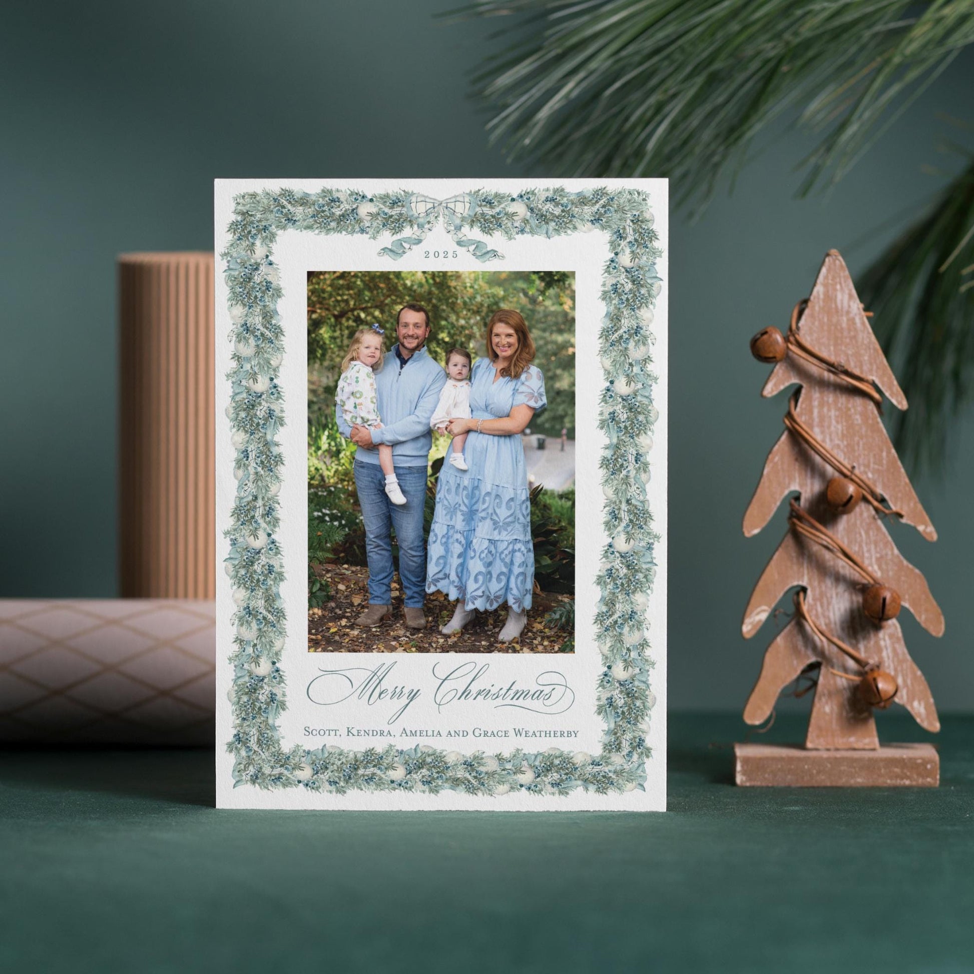 A Christmas card featuring a family photo of a man, woman, and child, with the text &quot;Merry Christmas Scott, Kendra, Amalia and Grace Weatherby&quot; displayed on the card. The card is placed next to a small wooden Christmas tree and a rolled-up paper, creating a festive holiday scene.