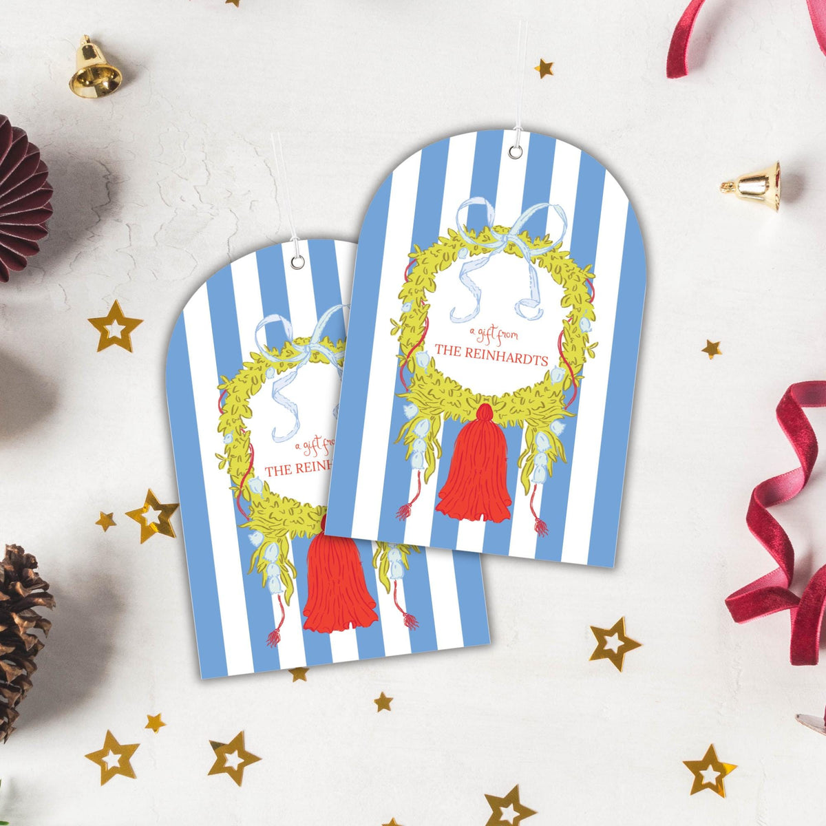 two blue and white striped gift tags with a wreath design and a red tassel, placed on a white background with Christmas decorations such as stars and pine cones.