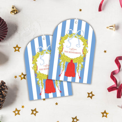 two blue and white striped gift tags with a wreath design and a red tassel, placed on a white background with Christmas decorations such as stars and pine cones.
