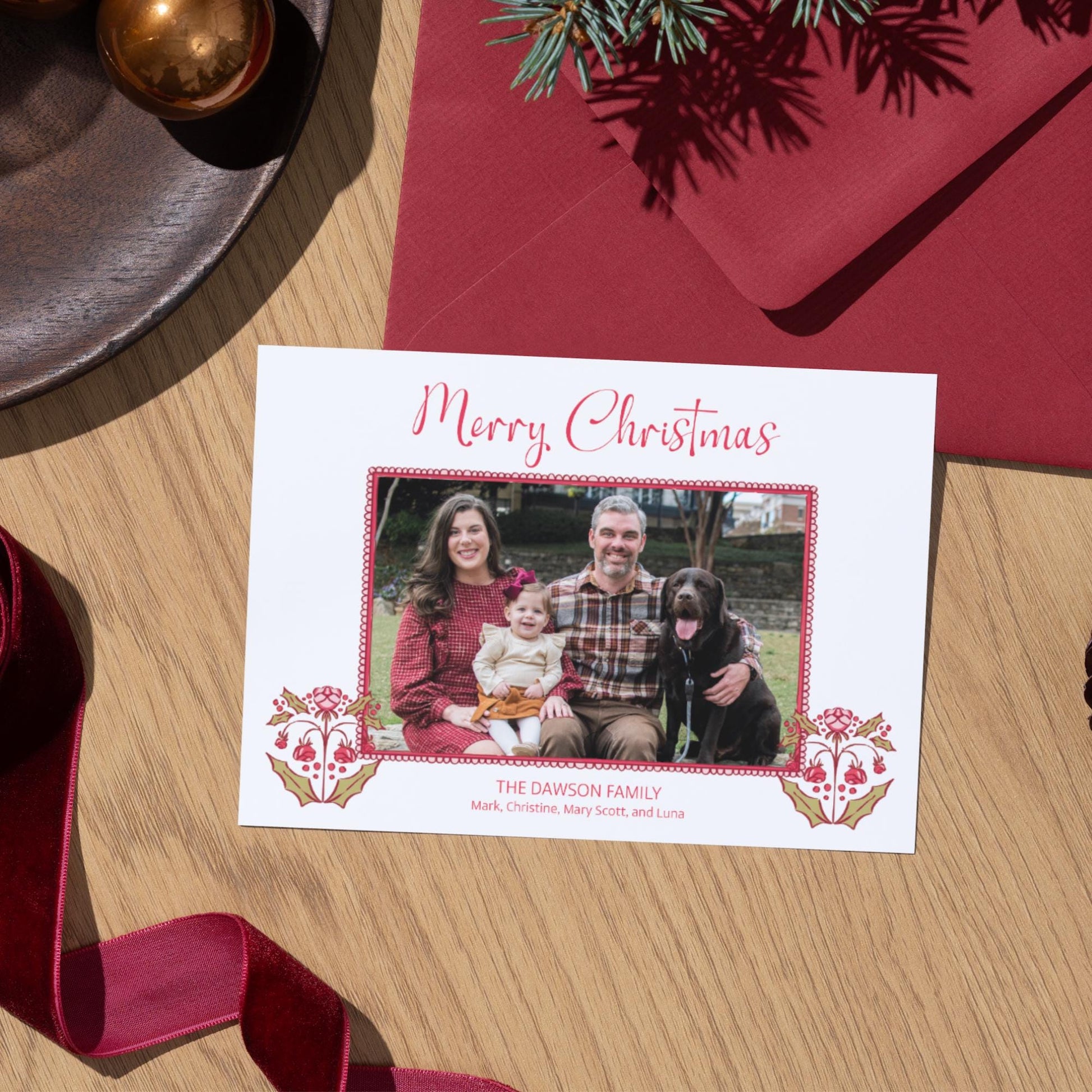 A Christmas card with a family photo of a man, woman, and child, along with a dog, sits on a wooden surface next to a red envelope and a red ribbon.