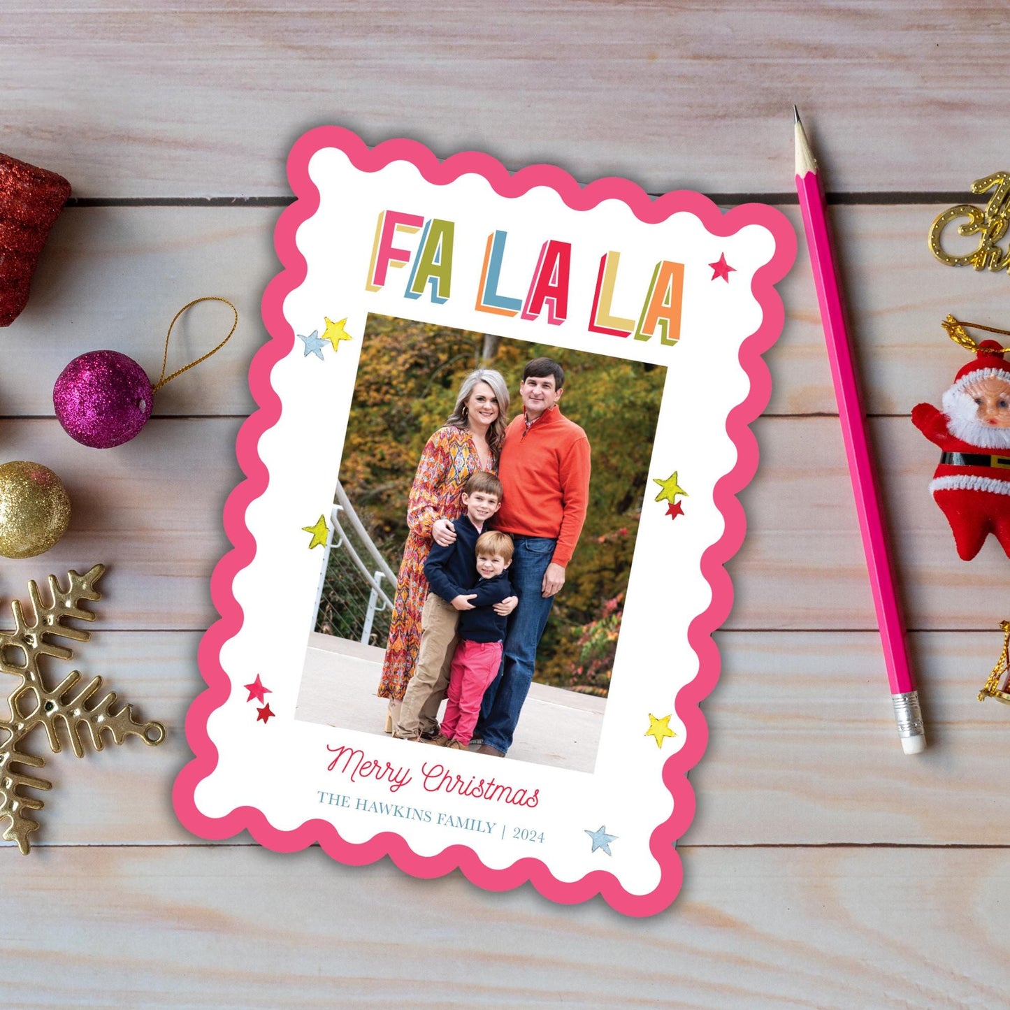 A family photo is displayed on a pink and white Christmas card, surrounded by various holiday decorations such as ornaments, a glittery ball, and a Santa Claus figurine.