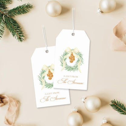 two white gift tags with illustrations of a wreath and a pine cone, placed on a beige background with Christmas ornaments and pine branches.