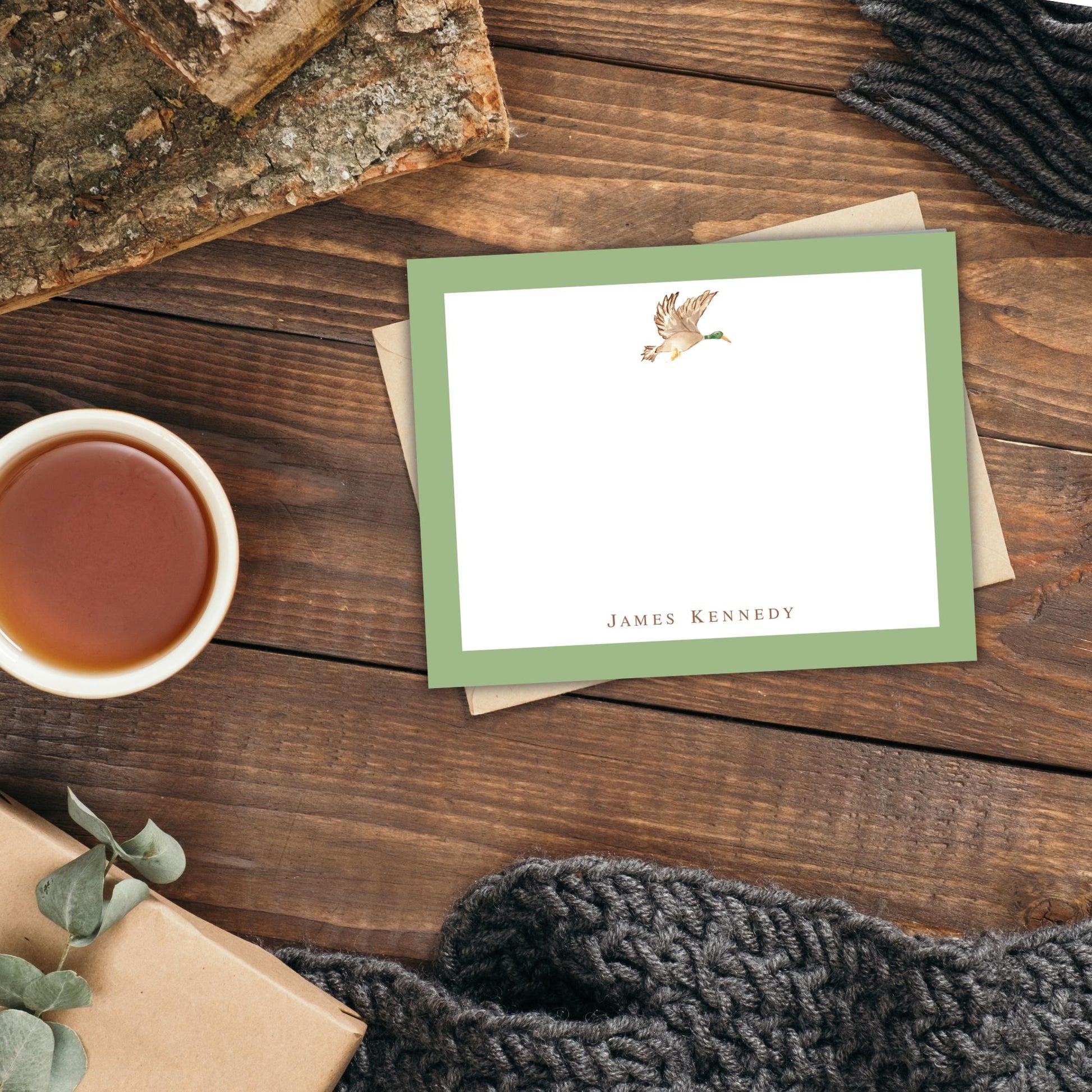 a wooden surface with a green-framed note card, a cup of tea, a wrapped gift, and a knitted blanket.