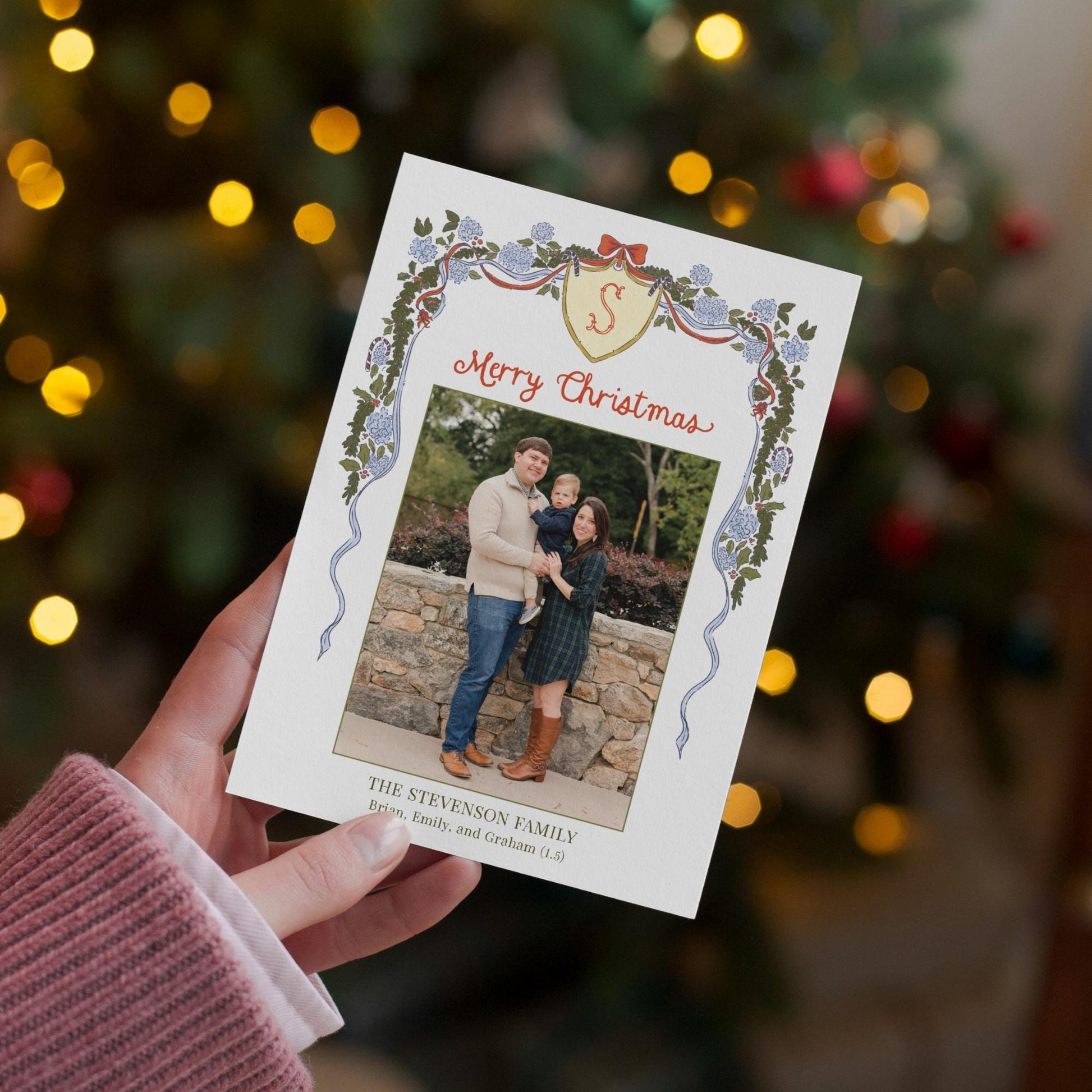 A hand holding a Christmas card with a family photo on it, set against a blurred background of a Christmas tree with lights.