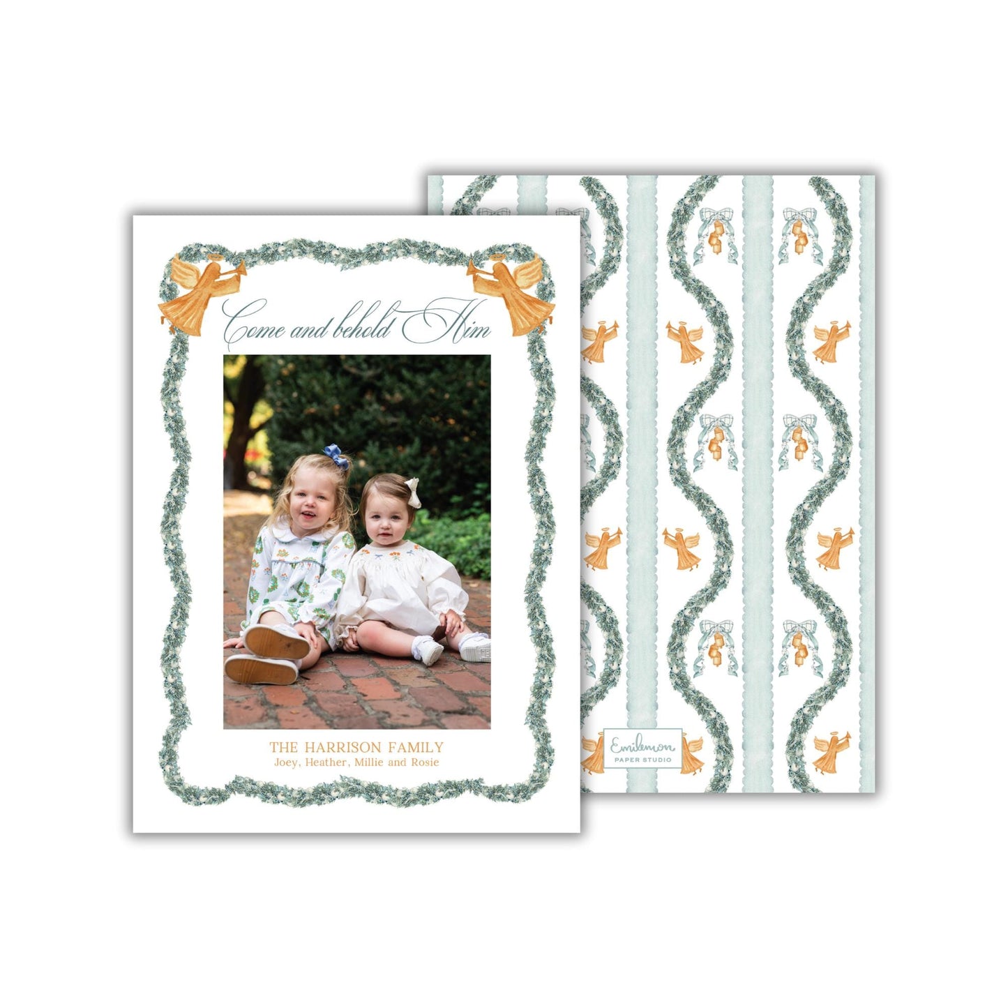 The image is a holiday card featuring two young girls sitting on a brick path, surrounded by a decorative border and a repeating pattern of angels and greenery.