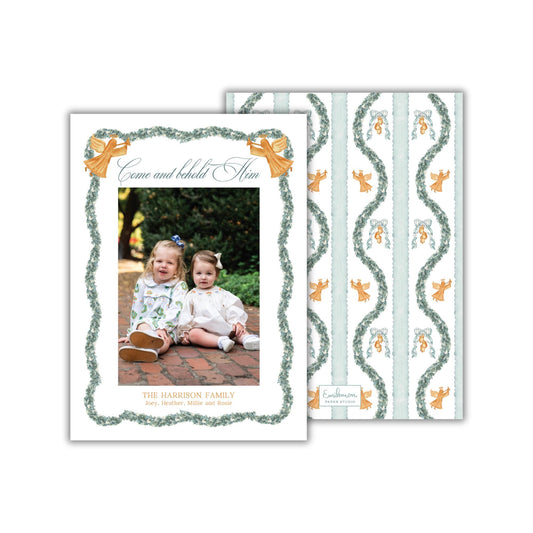 The image is a holiday card featuring two young girls sitting on a brick path, surrounded by a decorative border and a repeating pattern of angels and greenery.