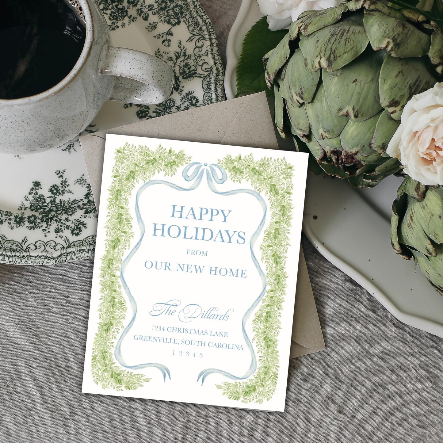 A white card with a festive holiday message sits on a table next to a teacup, a plate of flowers, and a green plant.
