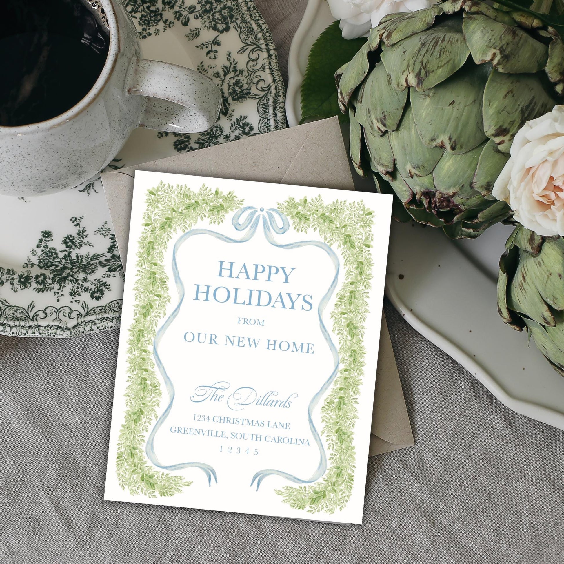 A white card with a festive holiday message sits on a table next to a teacup, a plate of flowers, and a green plant.