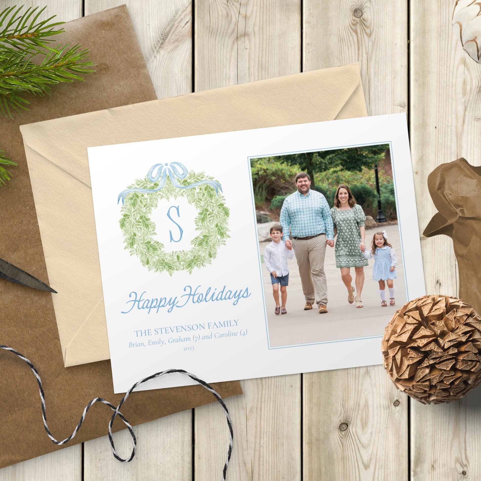 A family photo is displayed on a white card with a wreath design, surrounded by holiday decorations and a brown envelope on a wooden surface.