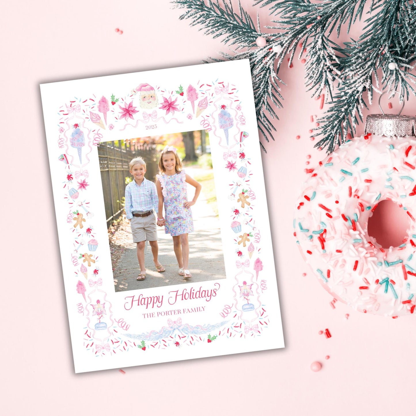 A white holiday card with a photo of two children, surrounded by a decorative border and text, placed next to a pink and white donut with sprinkles and a pine tree branch.
