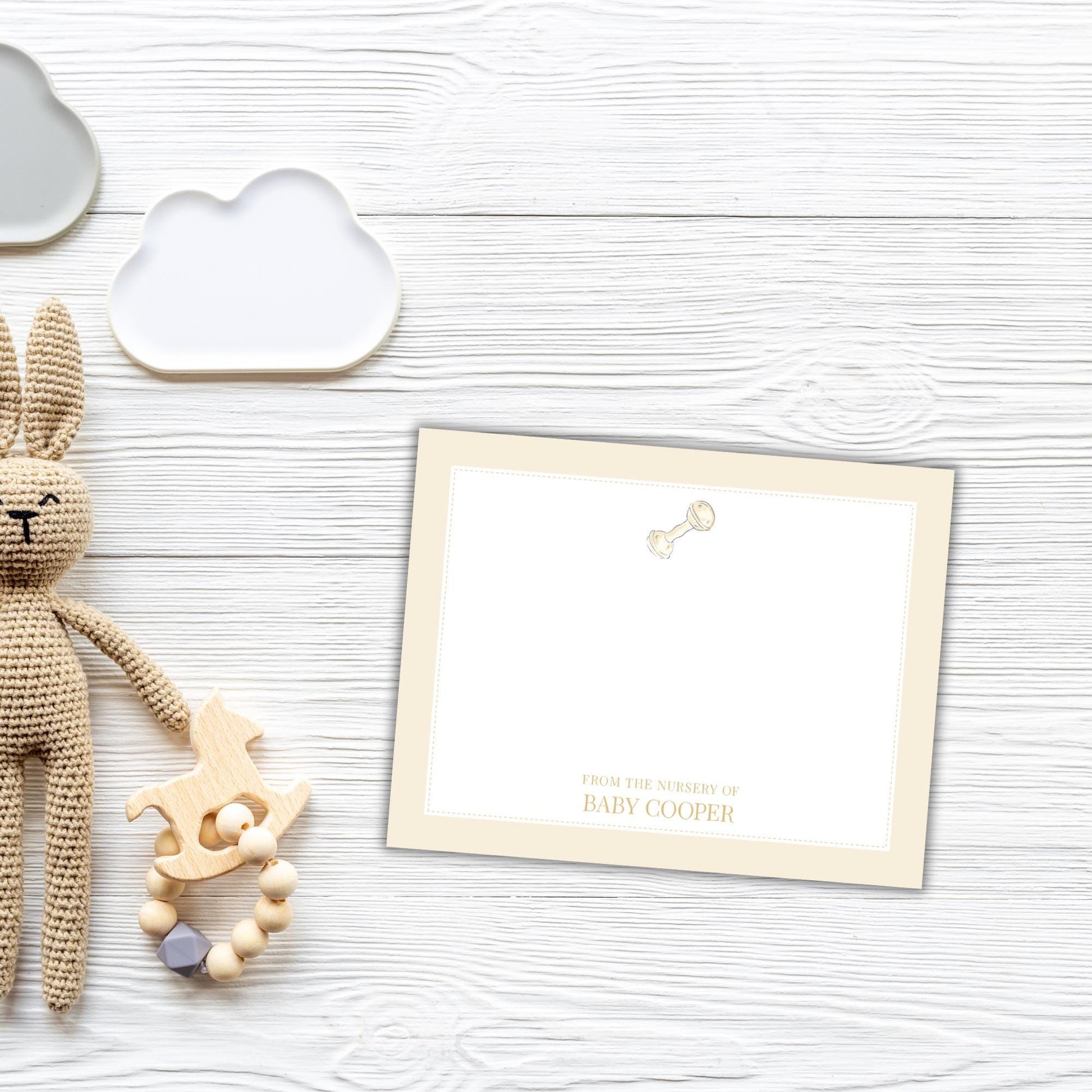 a white wooden surface with a crocheted rabbit toy, a cloud-shaped object, and a blank card with a baby rattle charm on it.