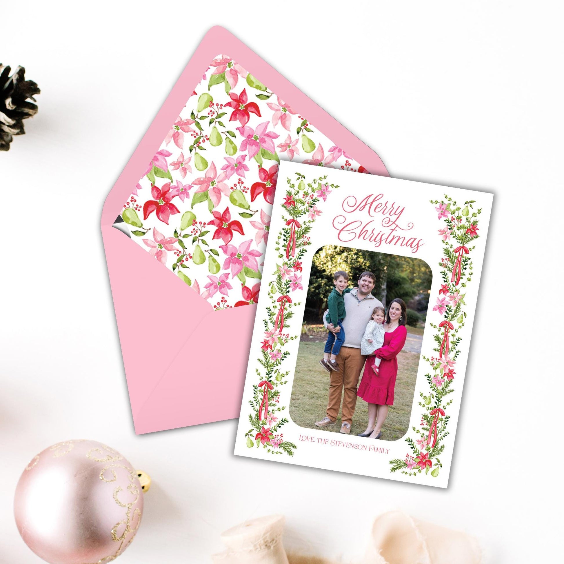 A pink envelope with a floral pattern and a Christmas card featuring a family photo, along with a pink ornament and a small tree branch, all placed on a white surface.