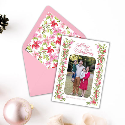 A pink envelope with a floral pattern and a Christmas card featuring a family photo, along with a pink ornament and a small tree branch, all placed on a white surface.