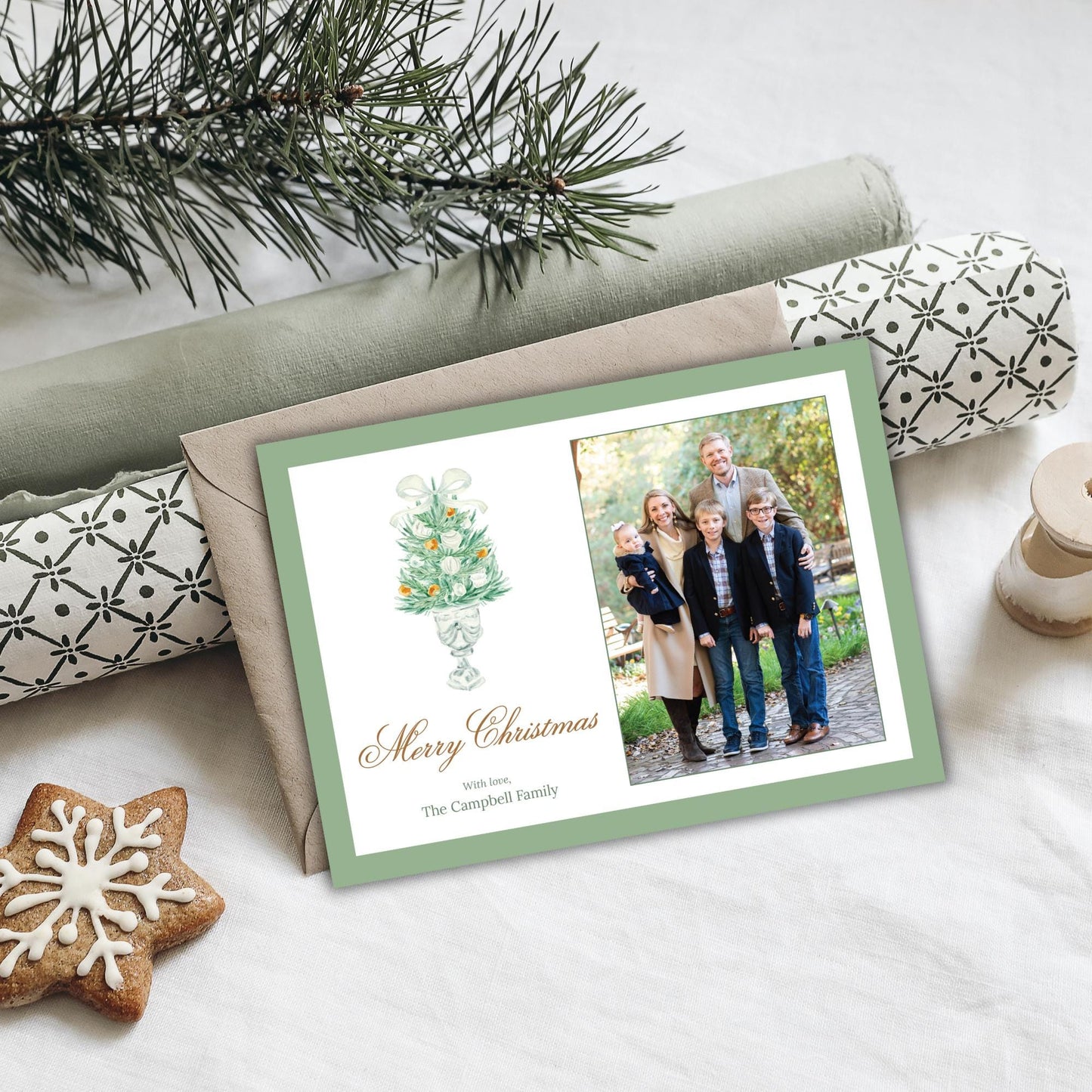 A Christmas card with a family photo and a Christmas tree illustration, placed on a table with a pine branch and a cookie.