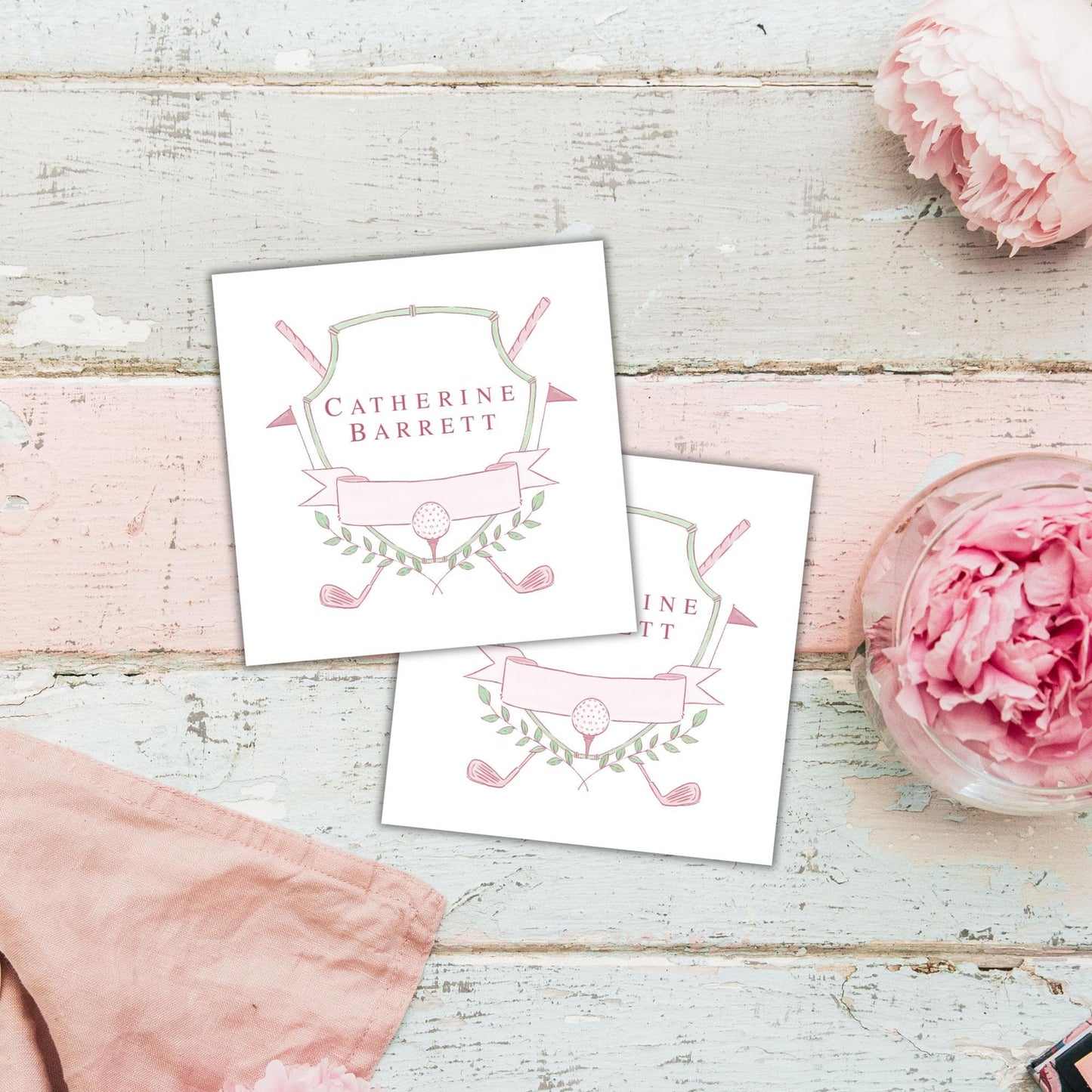 two white cards with a pink and green design, placed on a wooden surface alongside a pink flower and a pink cloth.