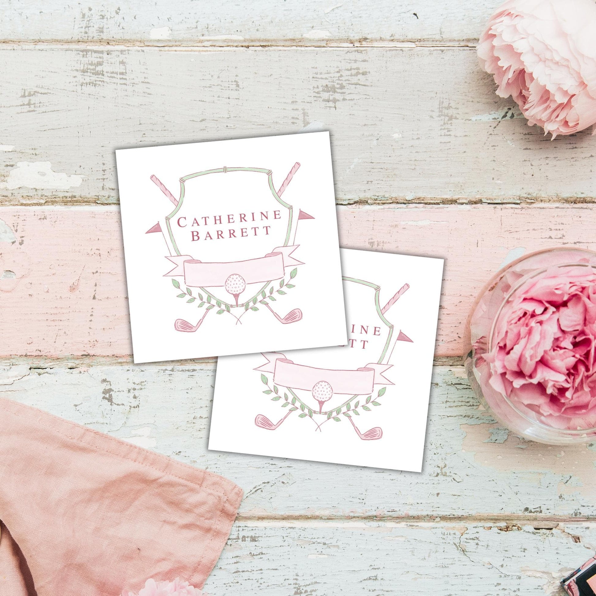 two white cards with a pink and green design, placed on a wooden surface alongside a pink flower and a pink cloth.