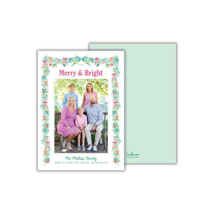 A family portrait of a group of people, including adults and children, sitting together in a garden setting. The text on the card reads "Merry & Bright" and "The Phillips Family" along with additional text that is too small to read clearly.