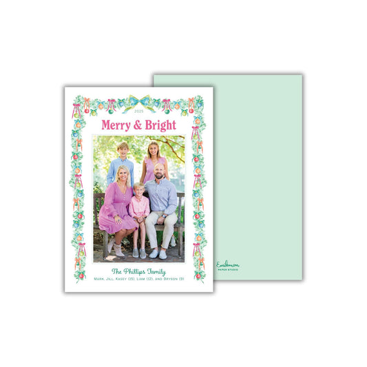 A family portrait of a group of people, including adults and children, sitting together in a garden setting. The text on the card reads &quot;Merry & Bright&quot; and &quot;The Phillips Family&quot; along with additional text that is too small to read clearly.