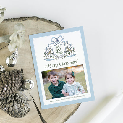 A Christmas card with a photo of two children, one boy and one girl, sitting together and smiling. The card has a blue background with a decorative border and the text "Merry Christmas?" and "Adam, Laura, Connor, and Anna Grace Kennedy" written below the photo.