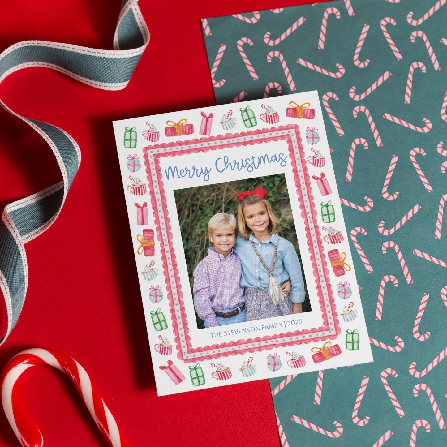 A Christmas card with a photo of two children is placed on a red surface, surrounded by candy canes and a patterned wrapping paper.