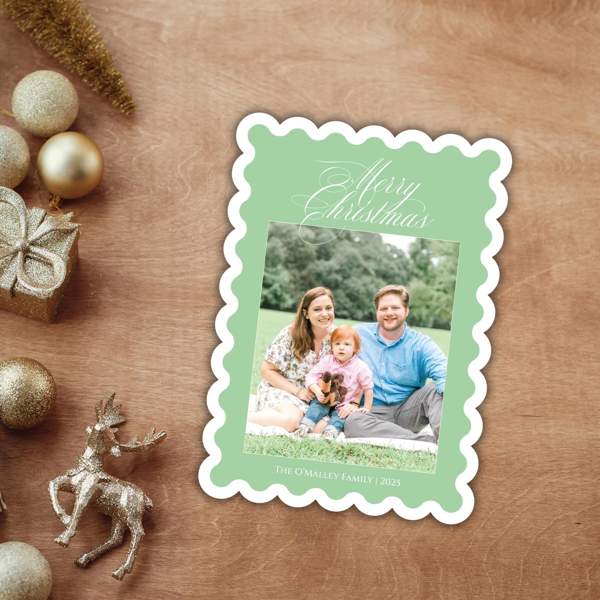 A family photo of a man, woman, and child is displayed on a green card with the text &quot;Merry Christmas&quot; and &quot;THE O&#39;NILLY FAMILY 2015&quot;. The card is placed on a wooden surface, surrounded by Christmas decorations such as ornaments and a small deer figurine.