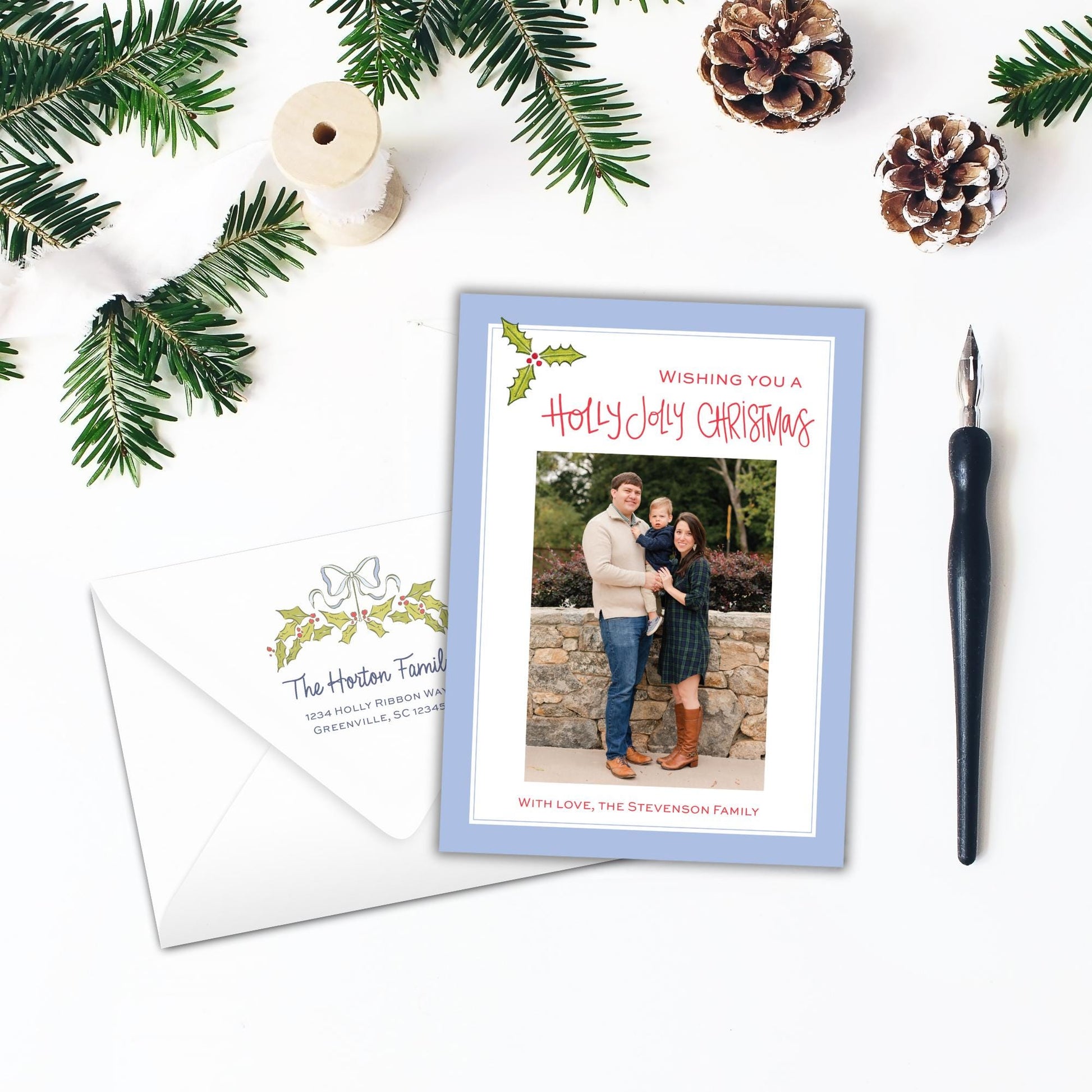 A holiday-themed card with a family photo, a pen, pine cones, and greenery on a white background.