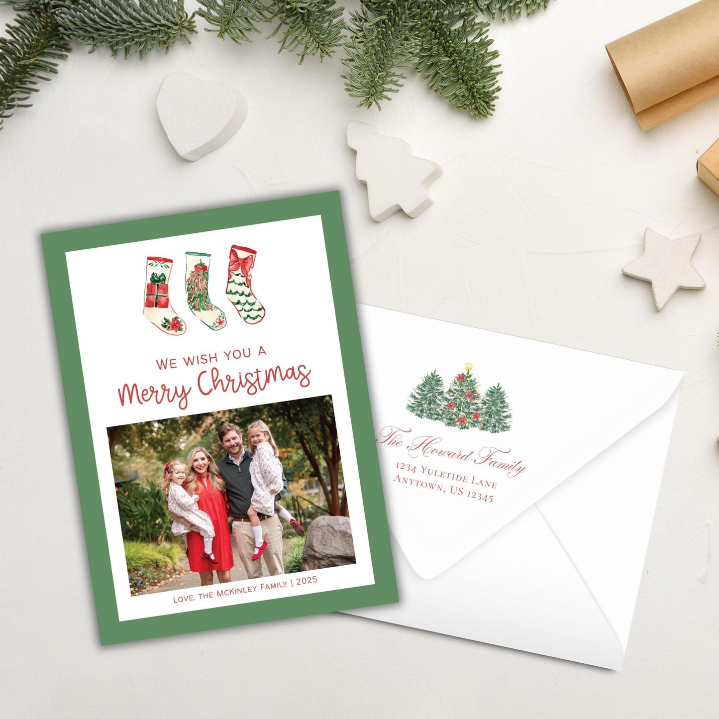 A green and white Christmas card with a photo of a family, along with the text &quot;We wish you a Merry Christmas&quot; and an address, is placed next to an envelope on a white surface.