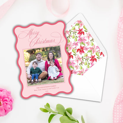 A pink Christmas card with a family photo of a man, woman, and two children, and a matching pink envelope with a floral pattern.