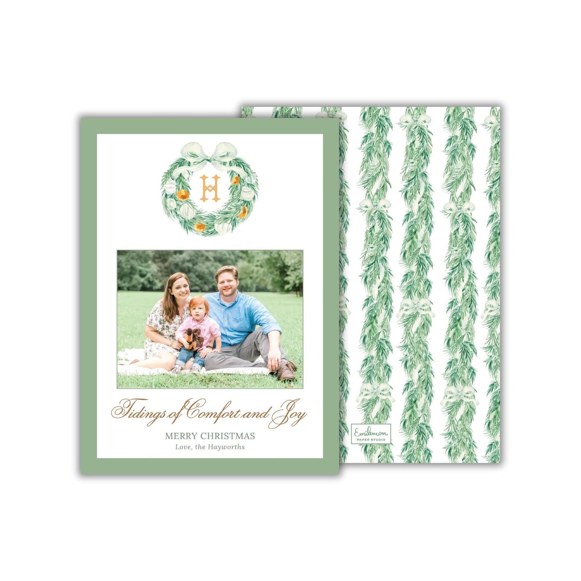 A family portrait of a man, woman, and child sitting together on a blanket in a grassy field, with a wreath above them.