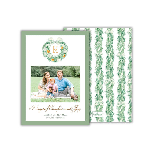 A family portrait of a man, woman, and child sitting together on a blanket in a grassy field, with a wreath above them.