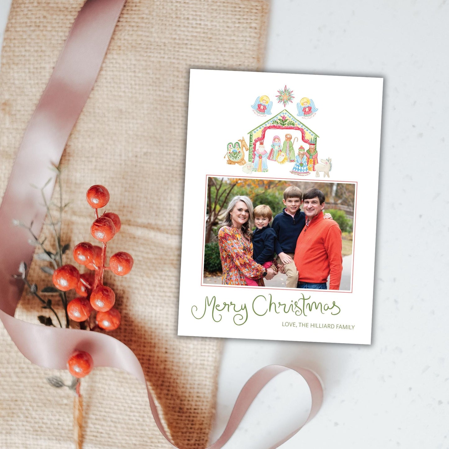 A Christmas card with a family photo and a nativity scene, along with a red berry decoration and a pink ribbon.