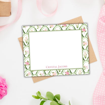 a blank card with a floral pattern and a nameplate, placed on a white surface alongside some pink flowers and a pink ribbon.