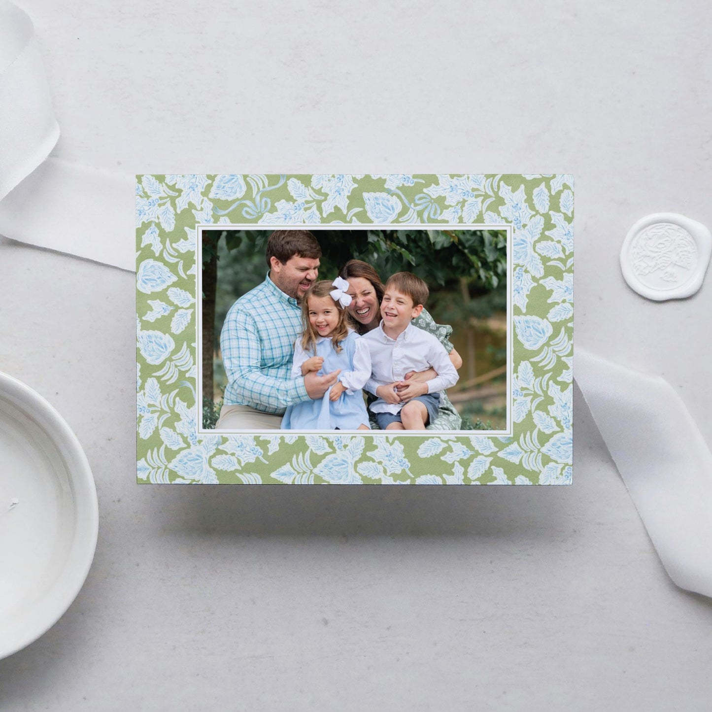 A family portrait of a man, woman, and two children, all smiling and embracing each other, is displayed in a green floral frame on a white surface.