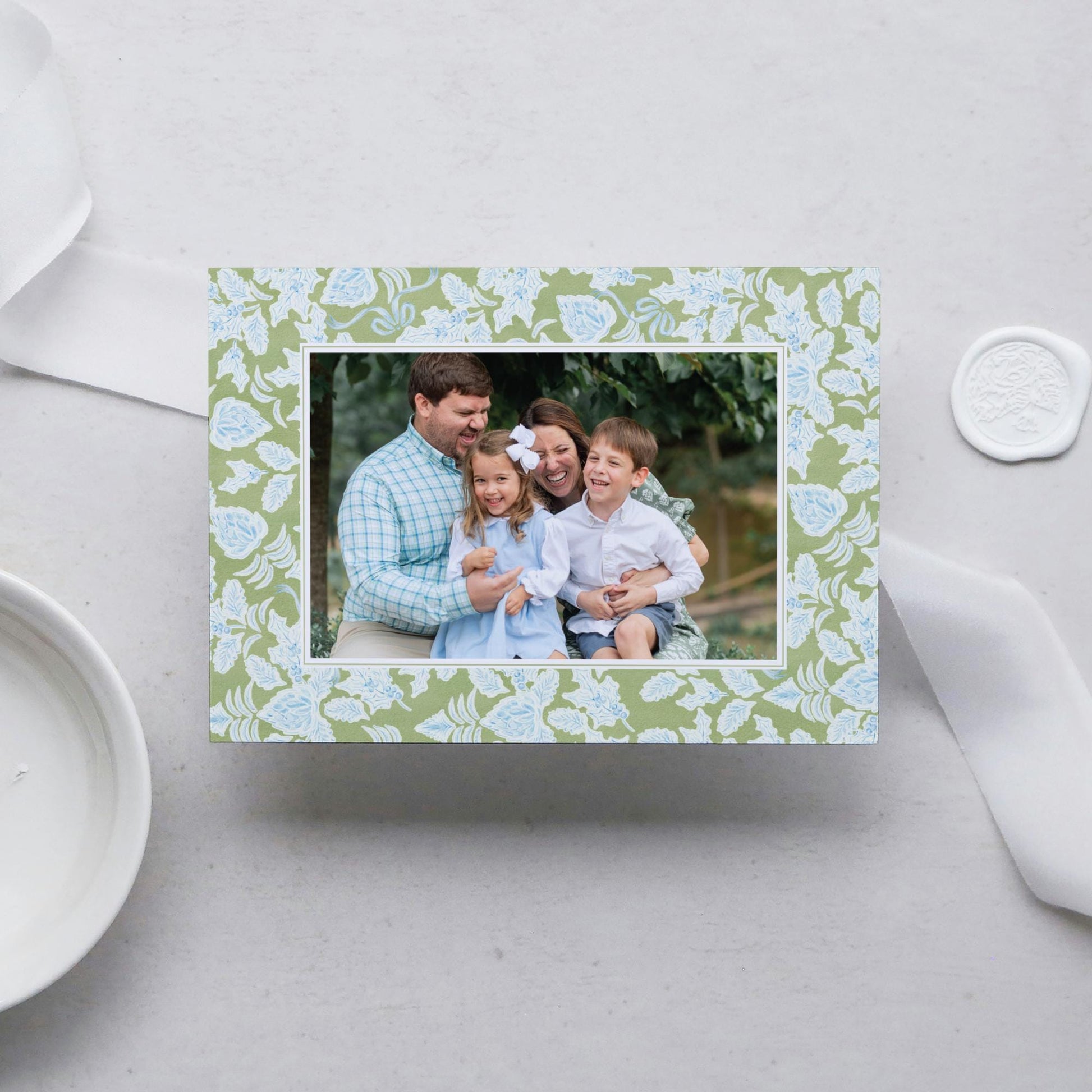 A family portrait of a man, woman, and two children, all smiling and embracing each other, is displayed in a green floral frame on a white surface.