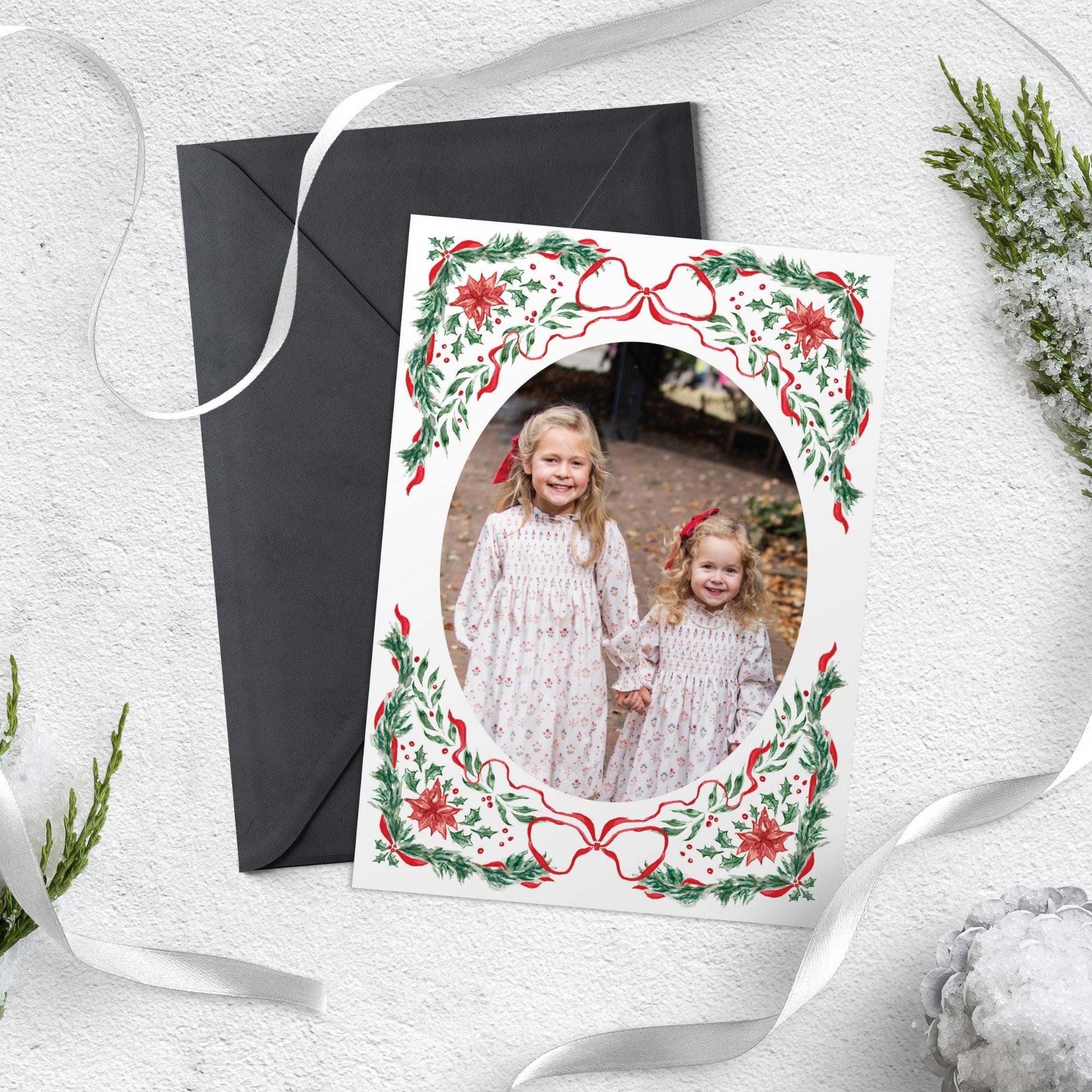 A Christmas card featuring two young girls in white dresses with red bows, surrounded by a decorative border of red and green leaves and flowers.