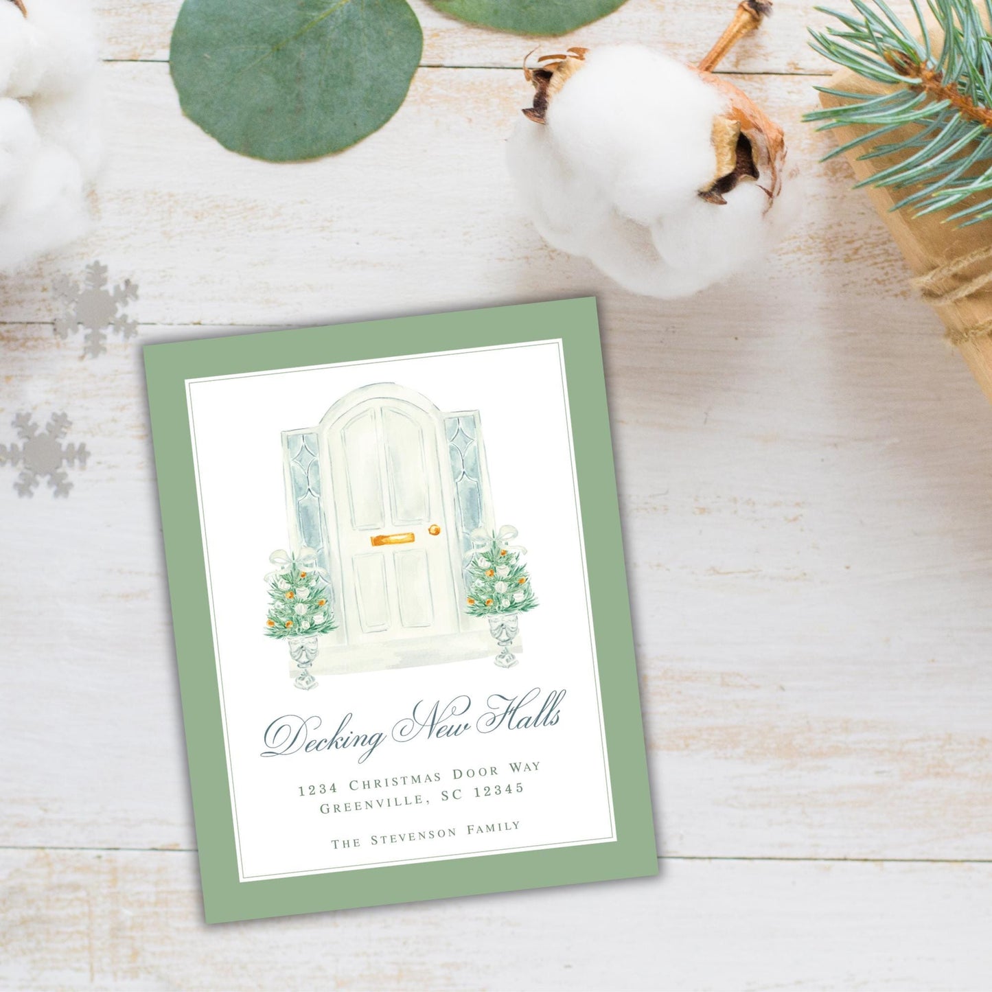 A green card with a white door illustration and text, placed on a wooden surface with a pine branch and cotton balls nearby.