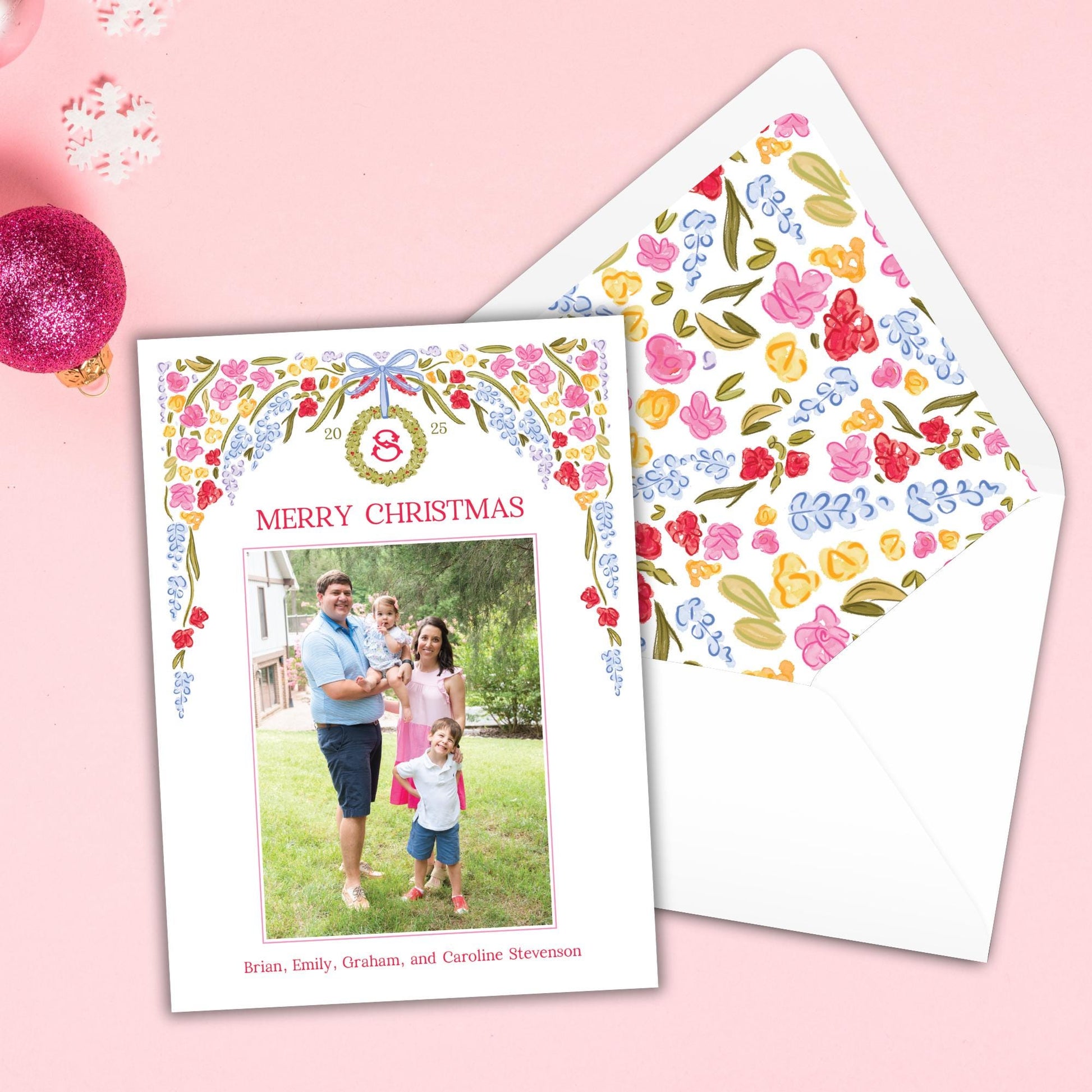 A Christmas card with a family photo and a floral pattern on the envelope is placed on a pink background.
