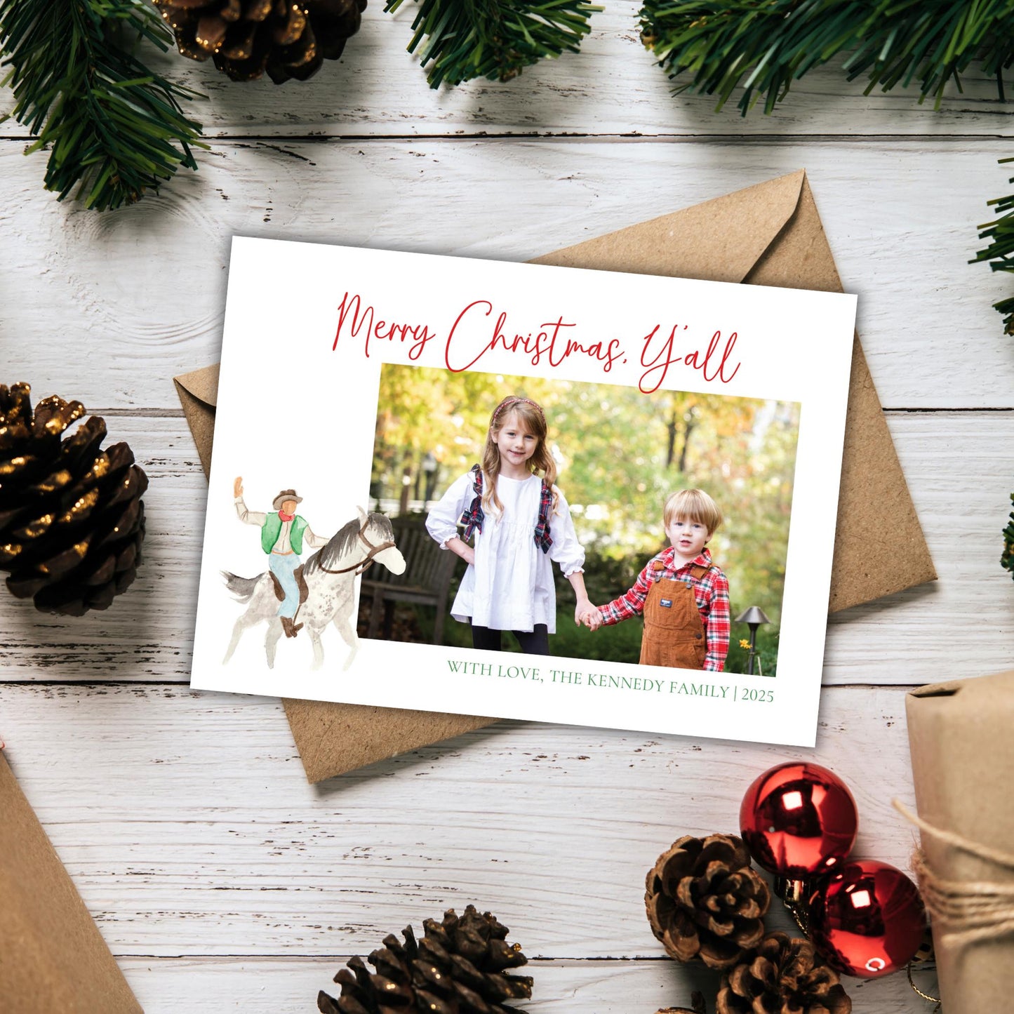 A Christmas card with a photo of two children, one riding a horse and the other holding hands, surrounded by pine cones and ornaments on a wooden surface.