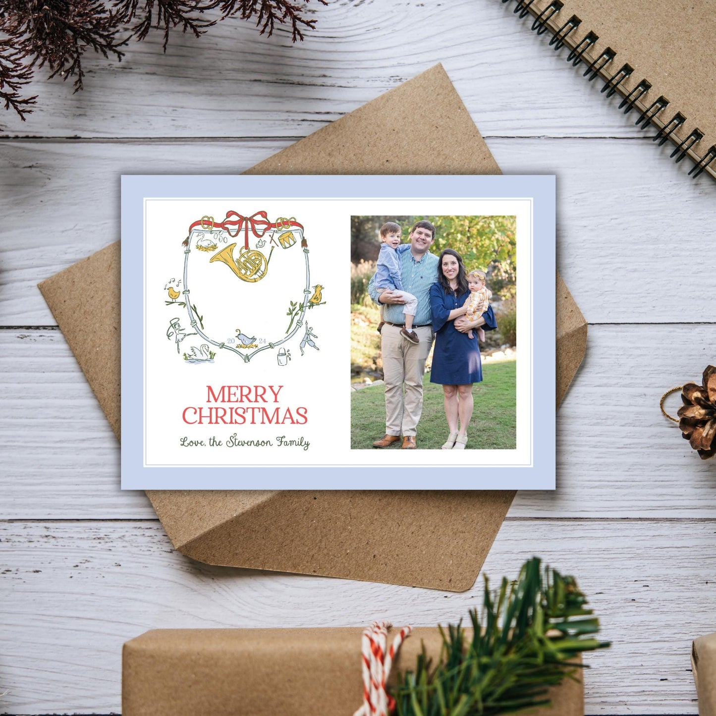 A family portrait of a man, woman, and child is displayed on a wooden surface, along with a greeting card that reads &quot;Merry Christmas Love the Swanson Family&quot;.